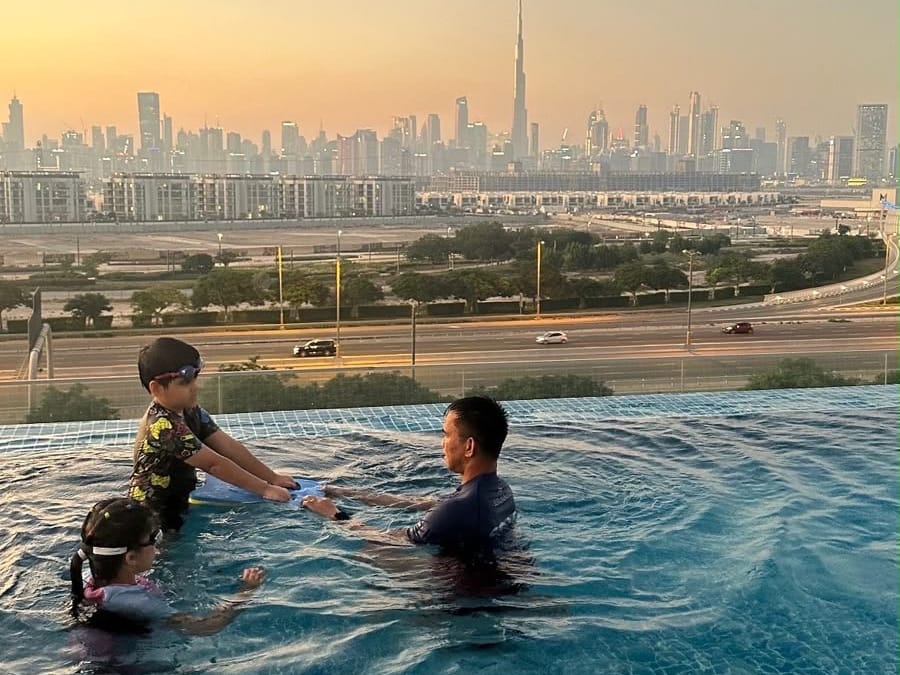 A coach guiding two children with a kickboard during a Wolf Kids swimming lesson, with the Dubai skyline behind