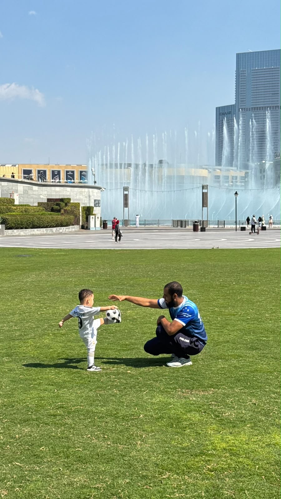 A Wolf Kids coach guiding one child's shot on a park lawn, the Dubai Fountain behind.